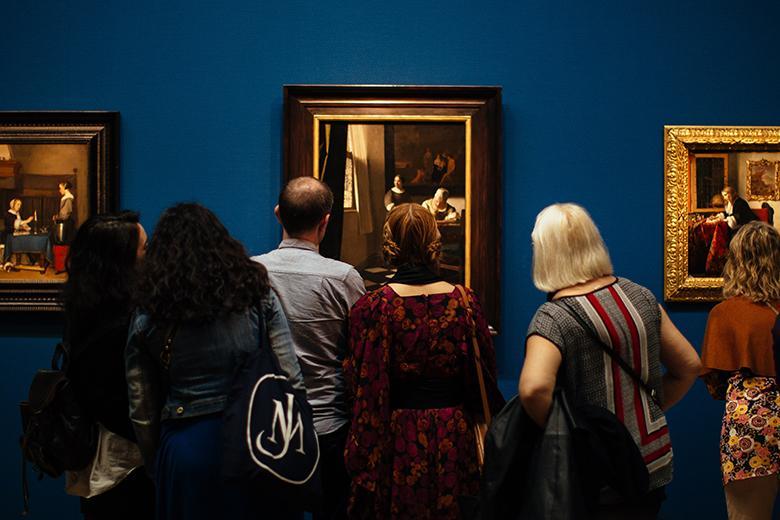 A photograph of people, with their backs to the camera, looking at three gilt-framed paintings on a blue wall.
