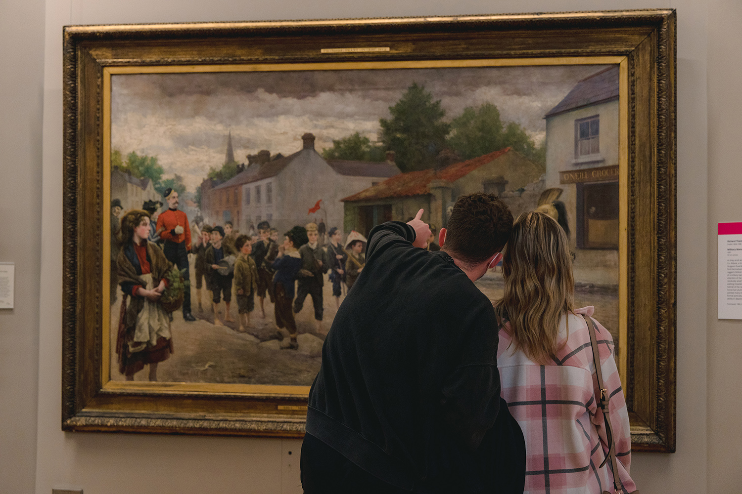 A photograph showing a man and woman standing looking at a large oil painting. Their heads are close together and the man is pointing at something in the painting.