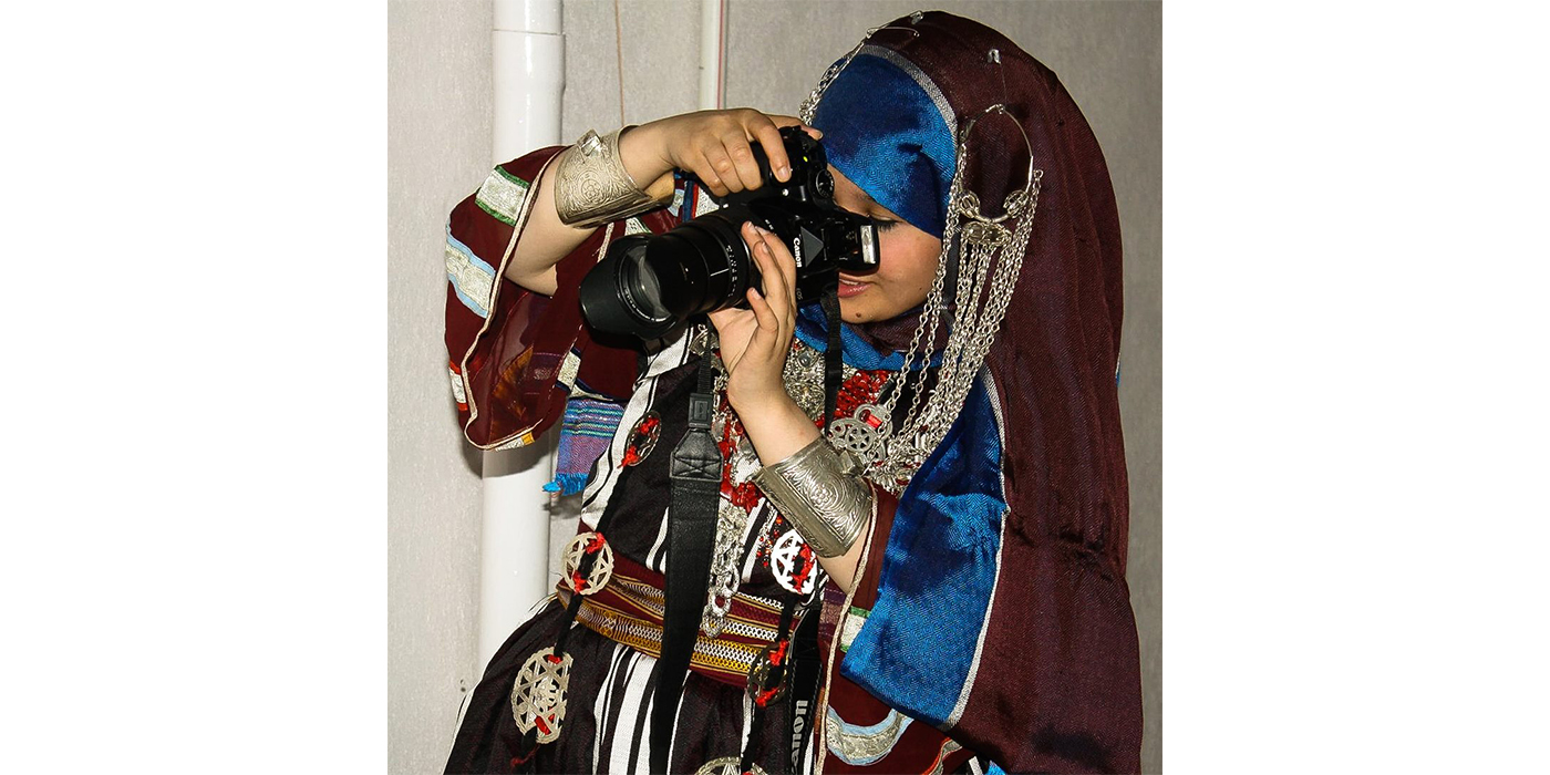 Portrait of a woman wearing a burgundy and blue headscarf and dress with decorative embroidery, as well as heavy silver cuff bracelets. She is using a large camera to take a photo.
