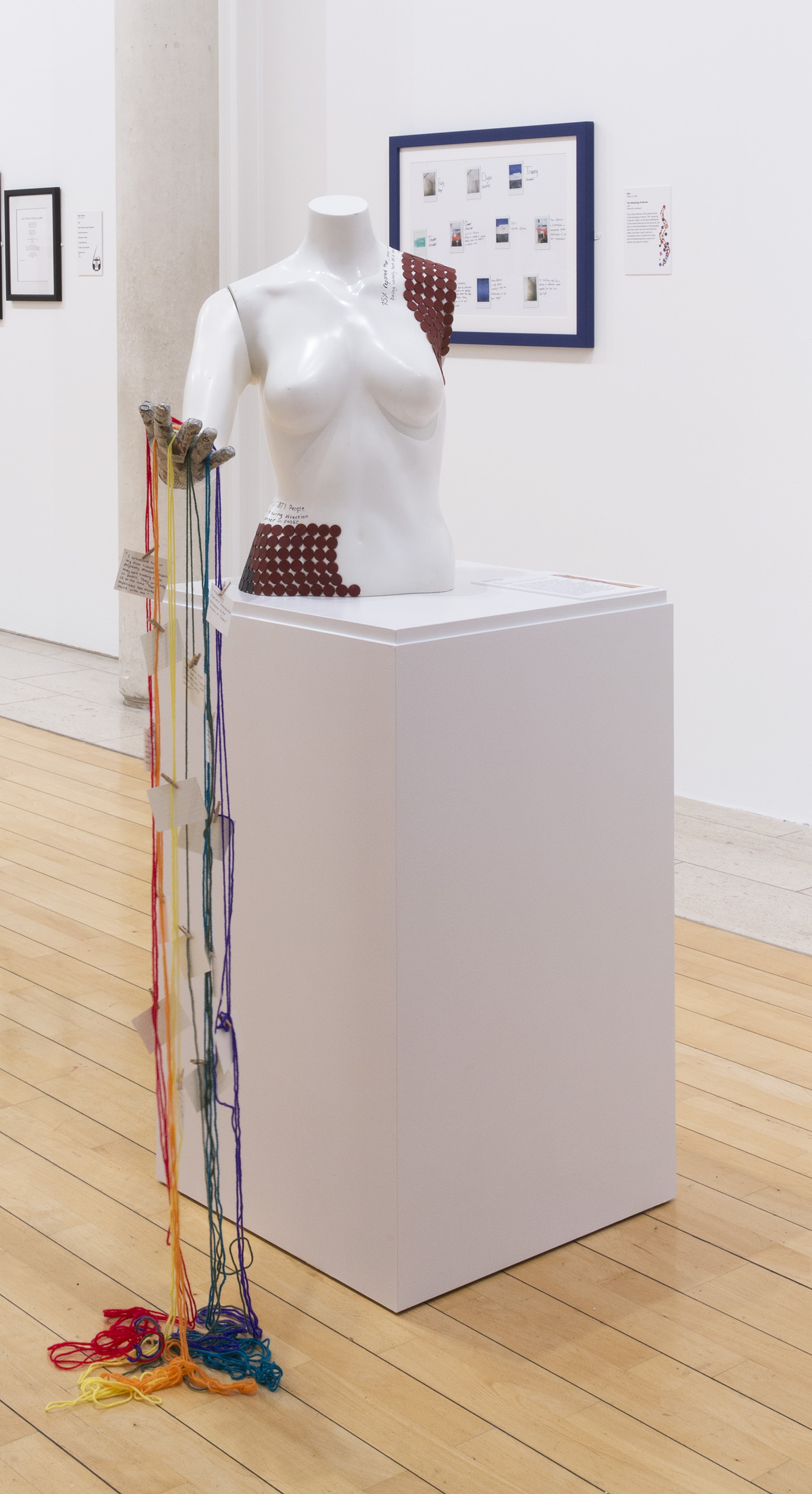 Installation shot of white bust on a white plinth with rainbow coloured strings trailing from the bust's hand to the floor