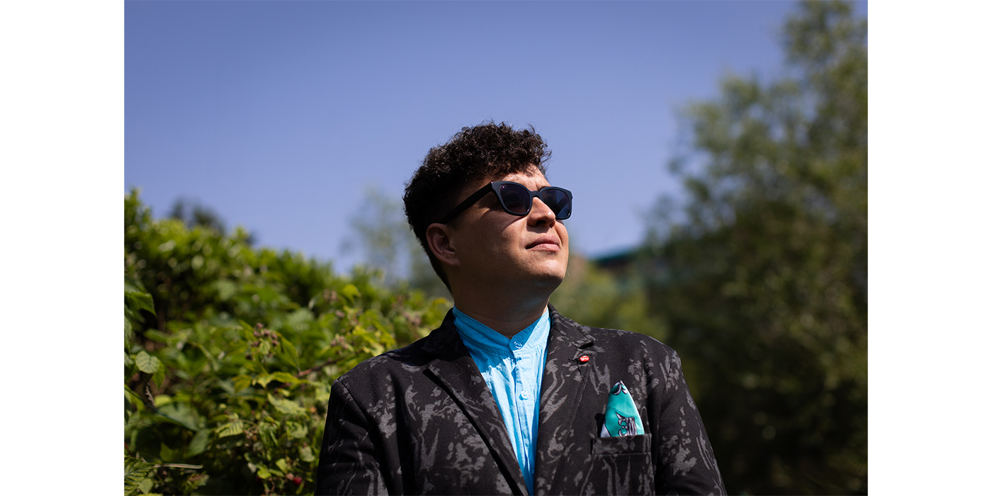 Photograph of a man with brown hair and a blue brocade jacket,  pictured looking up at a blue sky