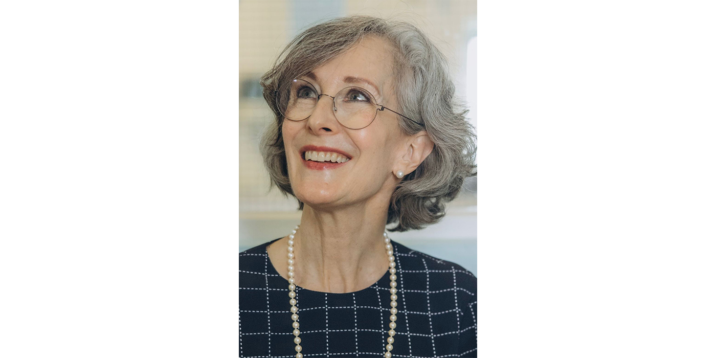 Headshot of a woman with grey curly hair and glasses, wearing a blue check top and a pearl necklace.