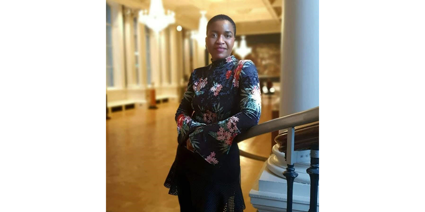 Portrait of a black woman wearing a floral velvet top in dark gem colours, pictured in a grand room with chandeliers and a large painting