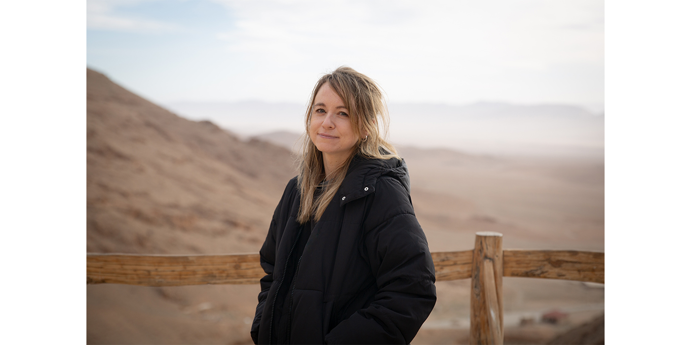 Photograph of a woman with blonde hair wearing a black jacket. She stands in a sandy desert landscape.