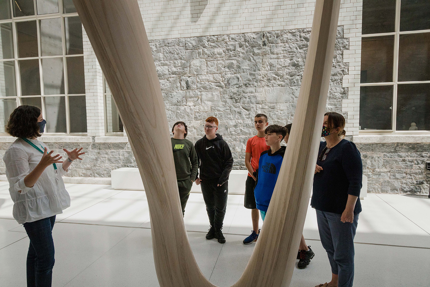 A Gallery guide speaks to a group of school students in front of a wooden sculpture