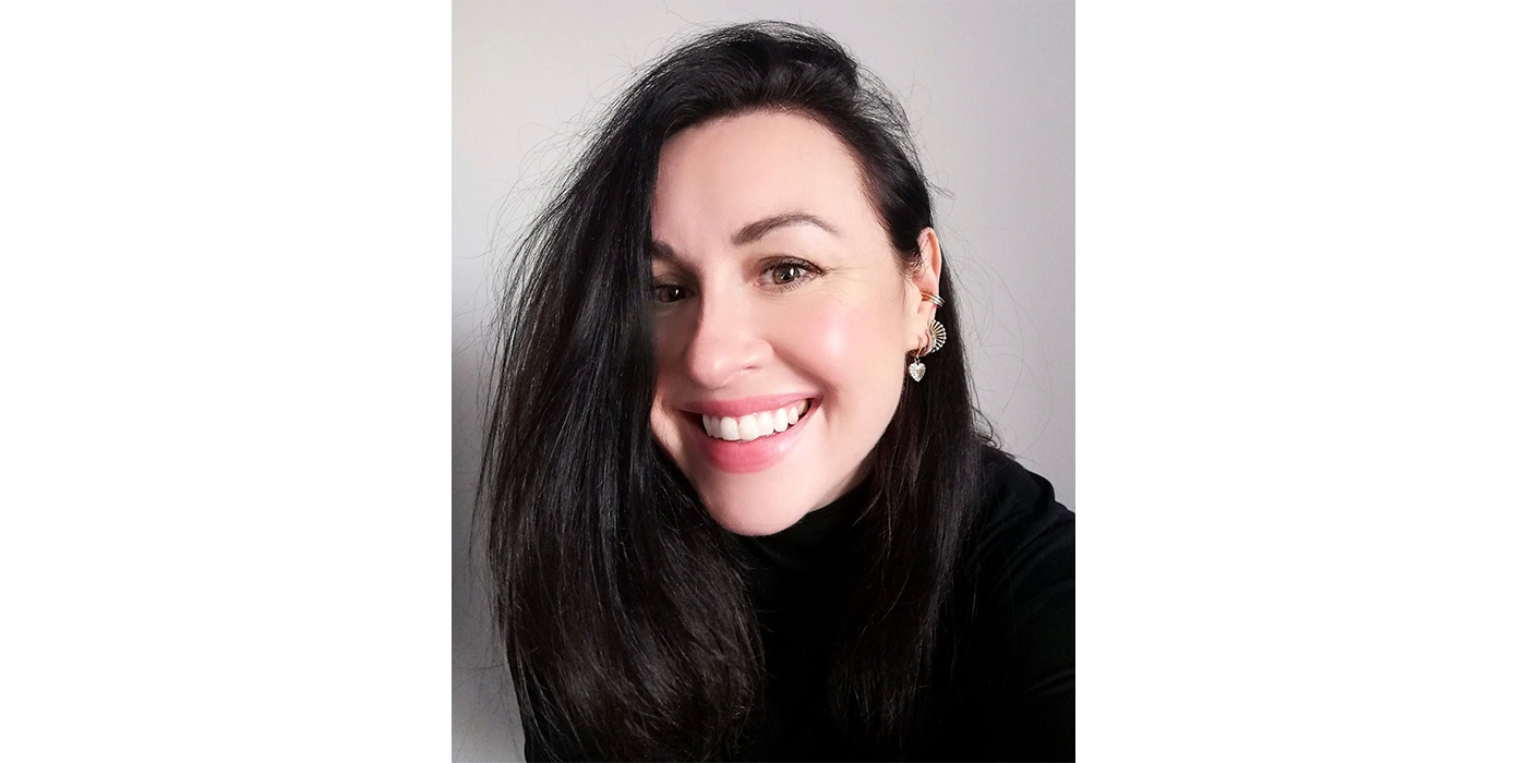 Headshot of a smiling woman with black hair, wearing a black top and a collection of silver earrings