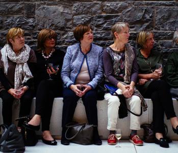 A group of people sit on a stone bench against an exposed stone wall. They appear to be listening intently to someone off-camera.