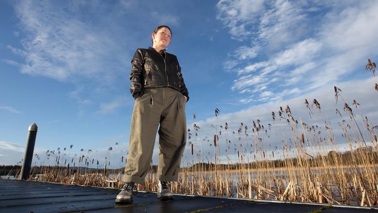 A photograph from ground level of a female figure wearing a black leather jacket standing by a lough.