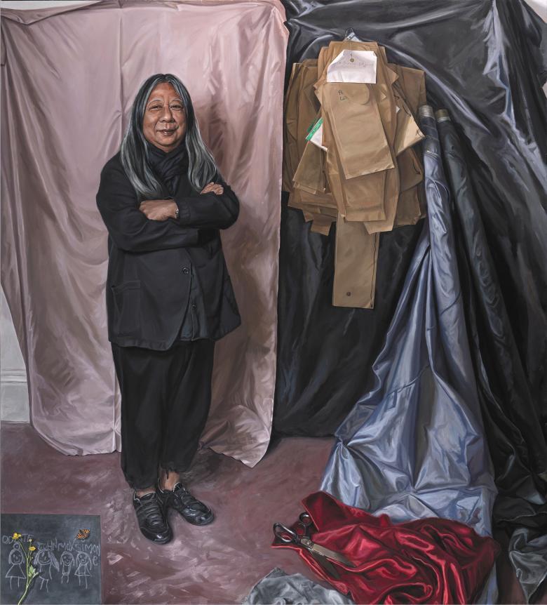 Painted full-length portrait of designer John Rocha wearing all black and standing in front of a wall covered in sheets of fabric and paper patterns.