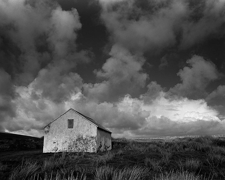 A black and white photograph with a low horizon and expansive cloudy sky, with simple whitewashed cottage standing in a field.