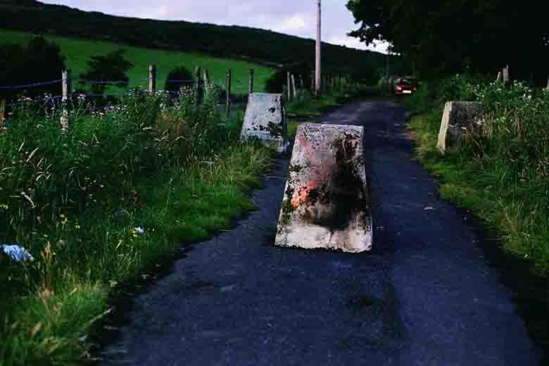 Photograph of a country road, bordered by green hedgerows and field, with three concrete bollards blocking the road.