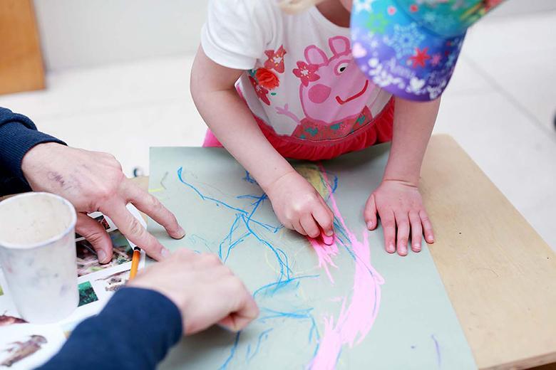 Photo of a young child drawing with an adult's hands stabilising the piece of paper.