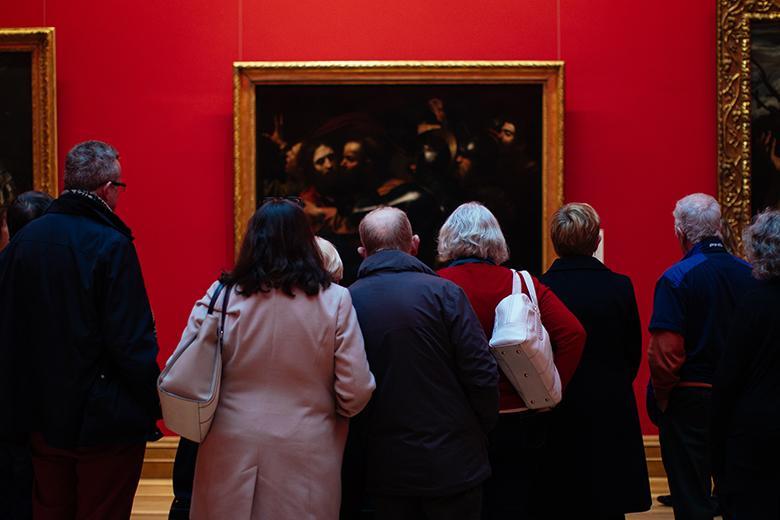 Photo of a group of people, with their backs to the camera, looking at Caravaggio's painting of the Taking of Christ, which is hanging on a red wall.