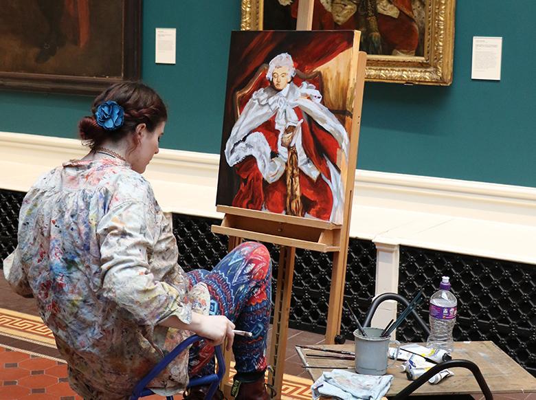 Photo of a young woman, dressed in paint-covered clothes, seated at an easel in a gallery and copying from a portrait.