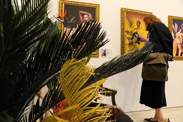 Photo of a woman looking at a framed photograph in an exhibition with plants in the left foreground.