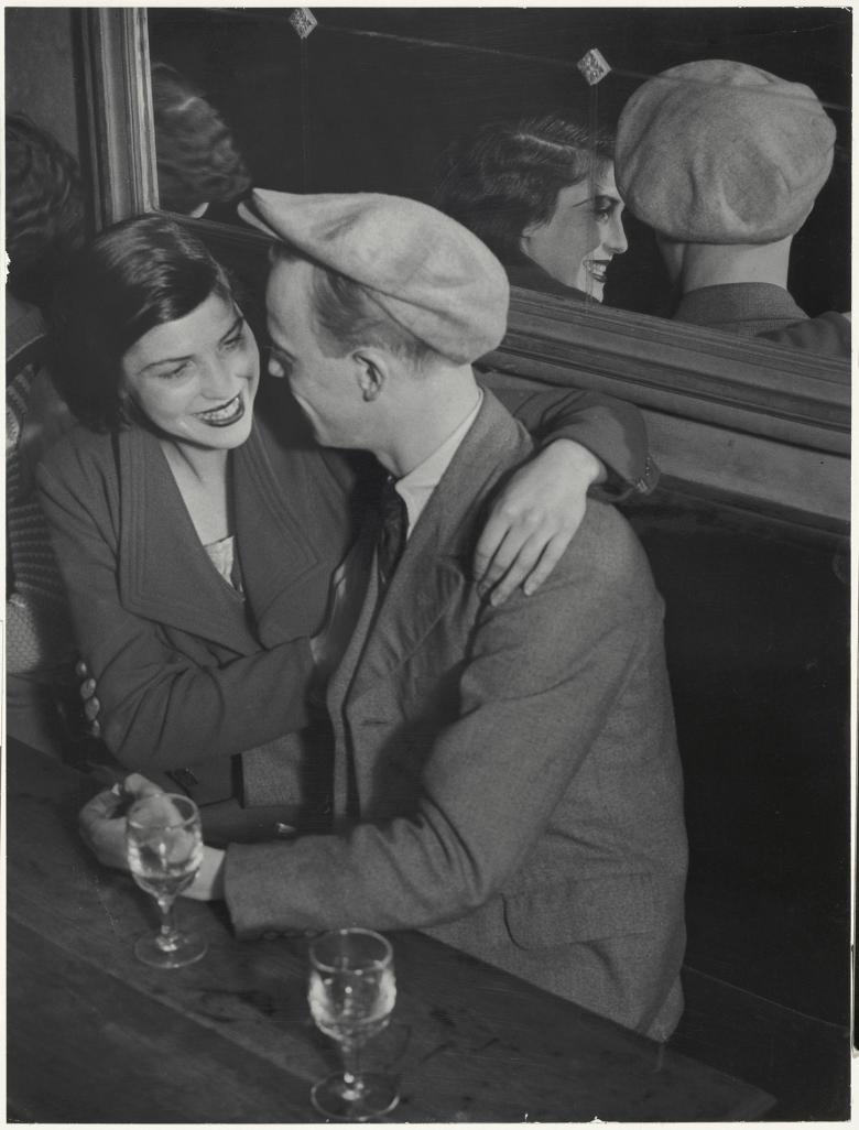 A black and white photograph of a woman and a man sitting side by side at a table, with two half full glasses of wine in front of them. They are turned towards each other, and the woman has her arm around the man's shoulder, and is smiling. Behind them is a large mirror in which we can see them reflected. 