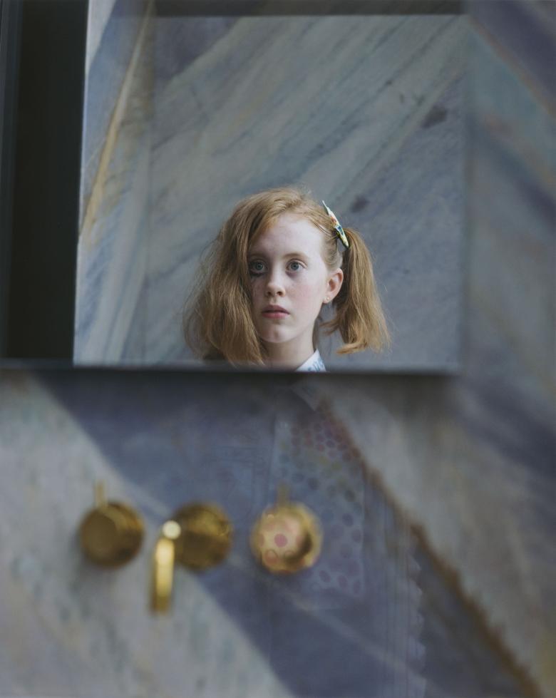 A photograph on paper. The head of a young girl is clearly visible, as though reflected in a square of mirror - on one side of her head her hair is pinned up in a ponytail, on the other side it is loose to her shoulders. One of her eyes has black markings under it. She is surrounded by abstract patterns - we can see her shirt faintly, as though reflected through a window. Out of focus beneath her face we see three brass fittings, like the fixtures of a bath.