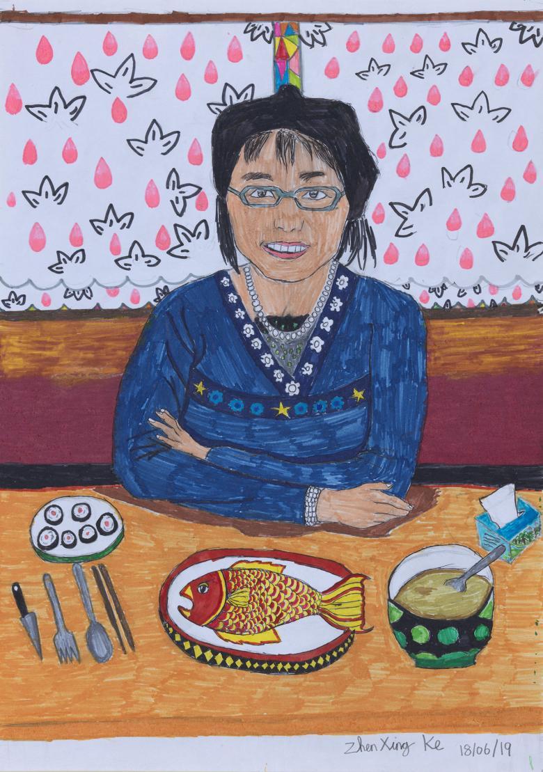 A woman in a blue top sits at a table with her arms folded and a smile on her face. In front of her, a table is laid for dinner, with a fish, a bowl of soup, and some californian rolls. Behind her, a pink white and black patterned wall. 