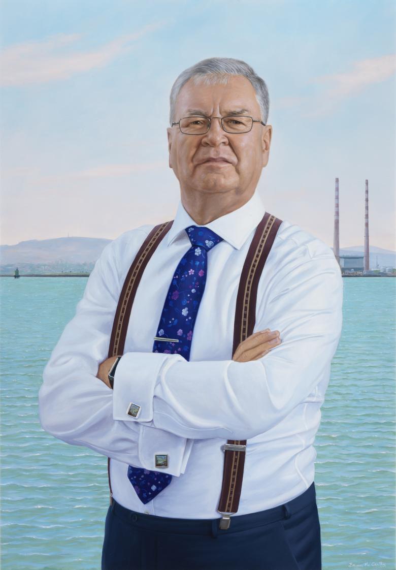 Three quarter length portrait of a man. He is wearing a white shirt, braces, and a blue tie. In the background we can see the sea, the mountains, and the Poolbeg towers.