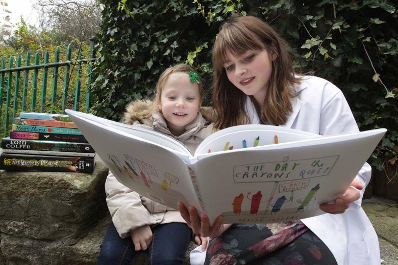 Photo of a woman and girl reading a children's book outdoors in a park.