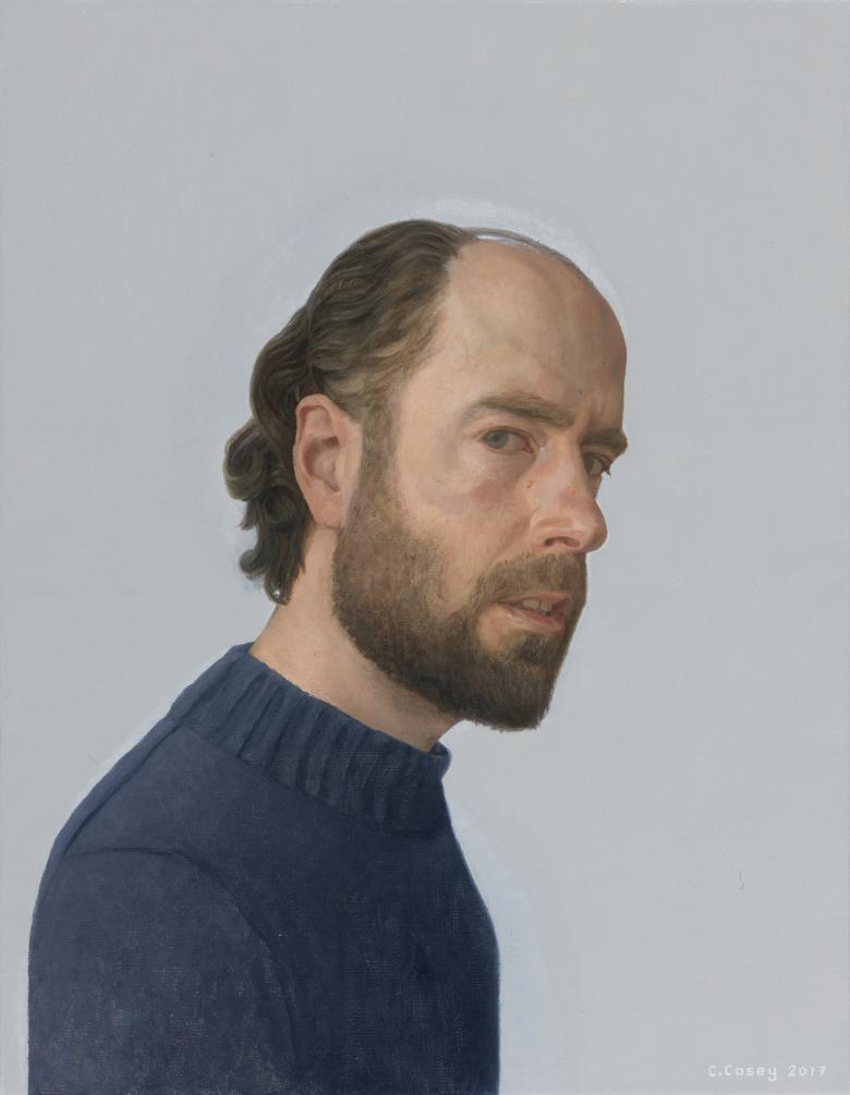 The subject of the portrait is standing side-on to the viewer, but his head is turned to look directly at us. He wears a navy woollen jumper.