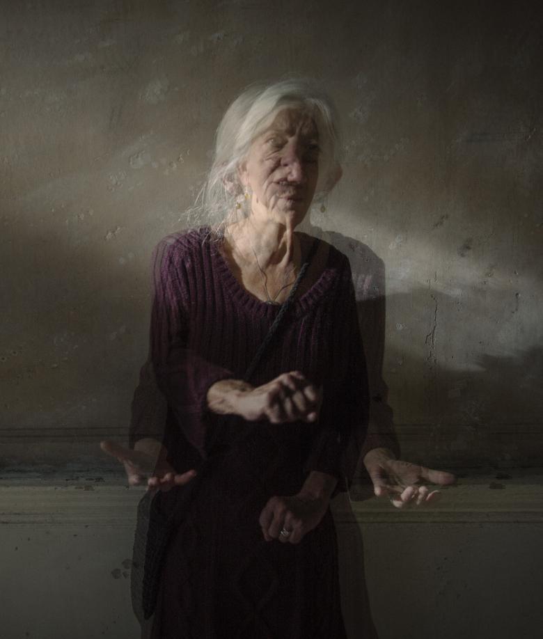This portrait has the effect of a moving image within a still image. The subject is sitting in a shaft of light in a dimly lit background. She appears very animated, with her hands and head in motion. 