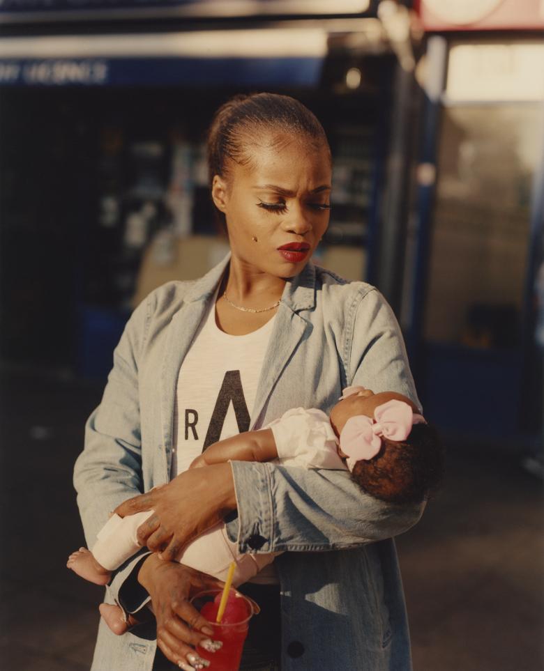 A woman dressed in a denim blazer and white t-shirt gazes down at the infant she cradles in her arms. The infant appears to be asleep, and has a large pink bow on her head. In the hand that is not holding the baby, the woman holds a red iced drink with a straw.