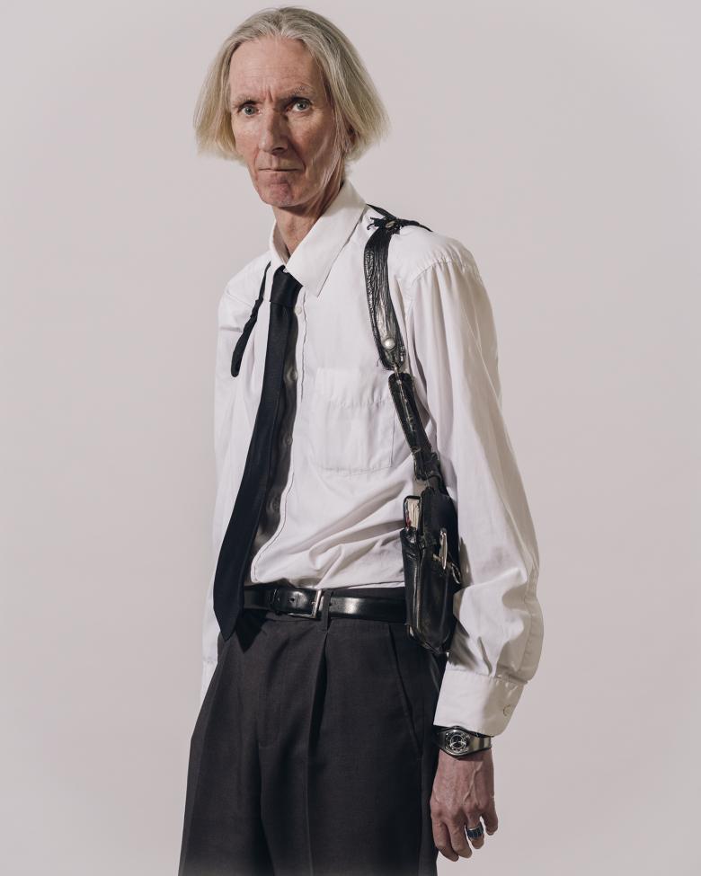 A three quarter length portrait of a male figure dressed in a white shirt, black tie, and back pants. He has white hair, and is looking directly at the viewer. He wears a black holster across his shoulders, which, when we look closely, holds a notebook and pen. 