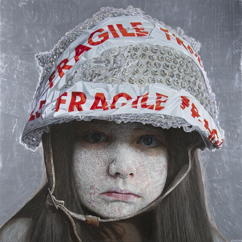A child stares directly at the viewer. Her face is coated in a white dust-like substance, and she wears a helmet on her head, fastened under her chin. The helmet is covered in bubble wrap, and has red and white tape on it with the word 'Fragile'.