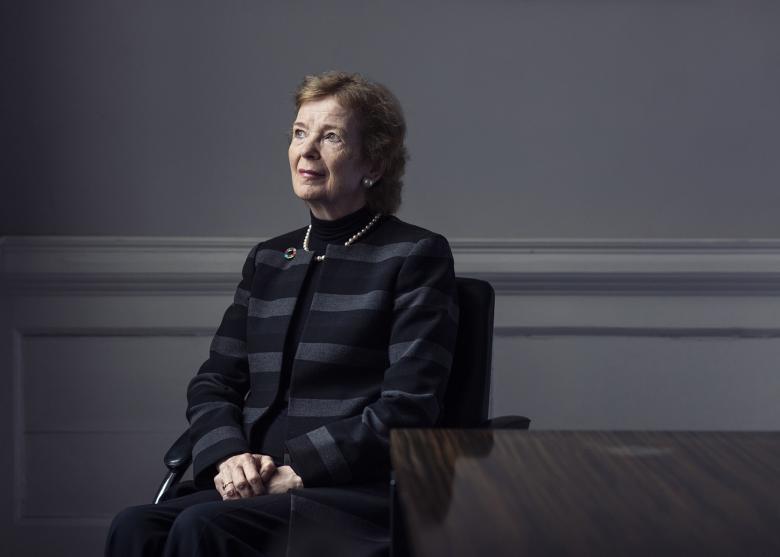 Former President of Ireland Mar Robinson sits beside a wooden table with her hands clasped in her lap. She is dressed in a grey and black striped jacket with a brooch at the lapel, and she gazes upwards and to her right.
