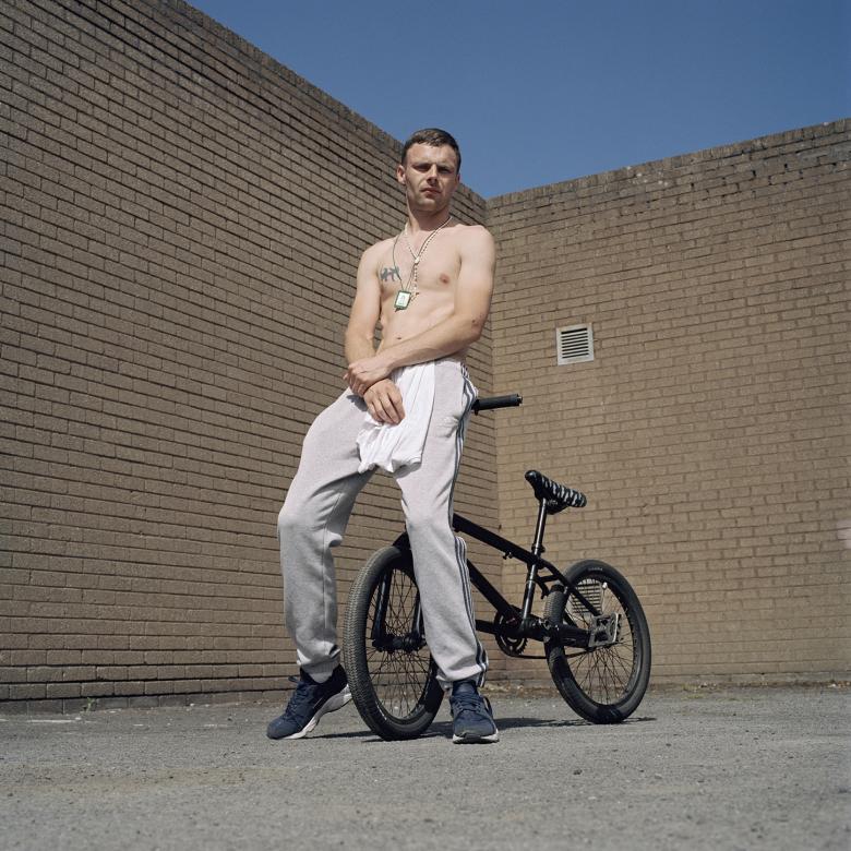 The subject of the portrait looks down a the camera lens, which is below him. He is leaning on the handlebars of a black bike which is behind him. He wears grey tracksuit pants and navy runners, and has a white tshirt tucked into his waistband. His hands are clasped in front of him, and he has a tatoo on his chest. Around his neck he wears a scapular and a rosary neads. Above his head, we see a section of blue sky above the brick wall behind him. 