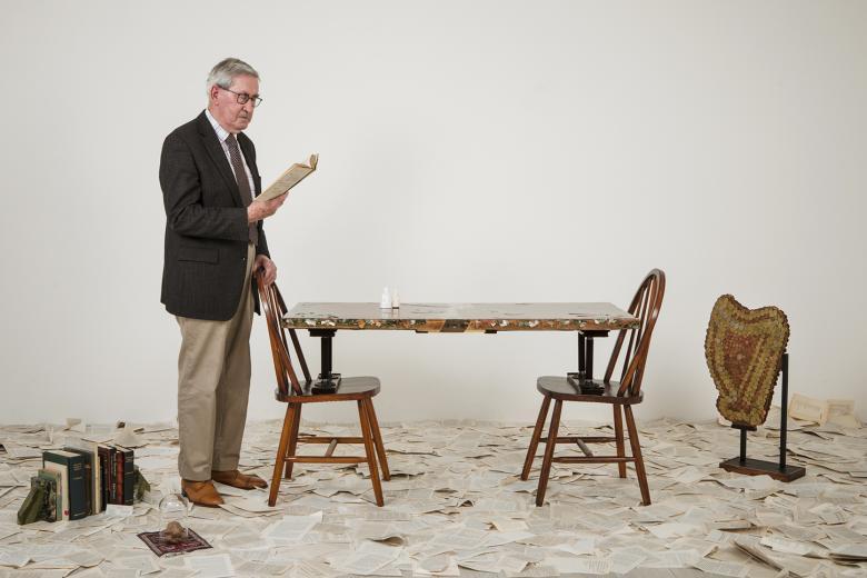 A man stands to the left of the frame wearing a suit. He is reading from a book, and he rests his hand on a chair in front of him. There is another chair opposite, and a table rests on the seat of the chairs. Strewn around the floor are pages from a book, and to the left is a small selection of books propped up between bookends. To the right is an efigy of a harp which appears to be made from coins. 