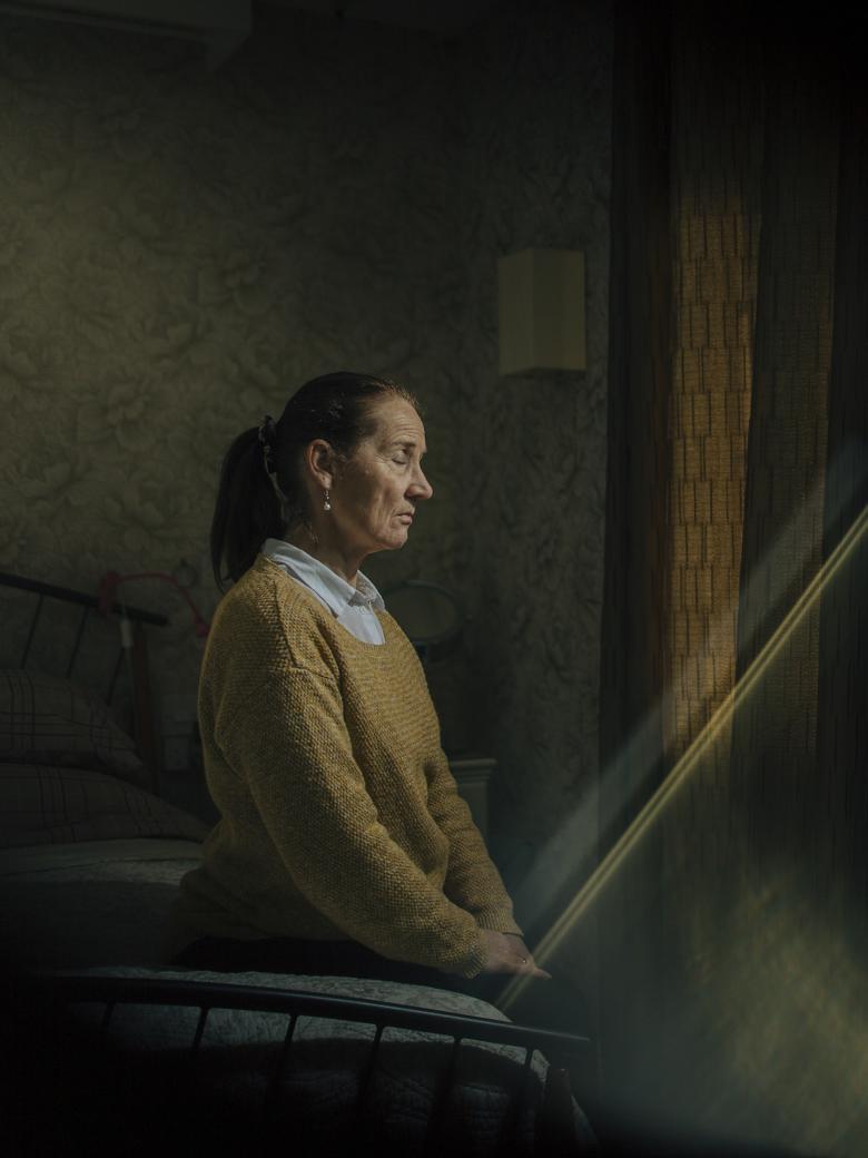 A woman in a mustard-coloured jumper over a white shirt sits on a bed with her eyes closed and her hands in her lap. She is turned towards a curtained window, and a shaft of light is coming into the darkened room in which she sits.