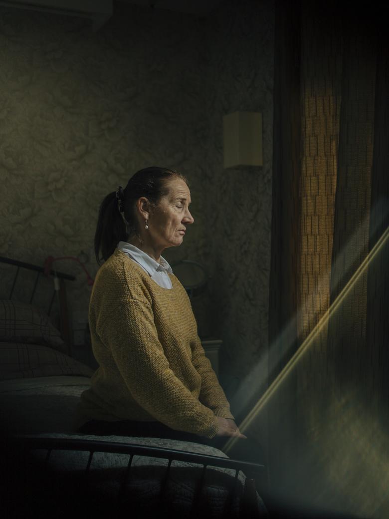 A woman in a mustard-coloured jumper over a white shirt sits on a bed with her eyes closed and her hands in her lap. She is turned towards a curtained window, and a shaft of light is coming into the darkened room in which she sits. 