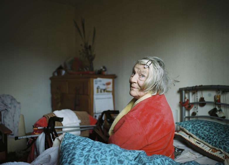 A woman in a red and yellow dressing gown sits on an unmade bed with a blue cover, looking at the viewer over her shoulder with an amused expression on her face. In the background we see a chest of drawers with a calendar attached to the side, and flowers and other belongings on top. The room is filled with signs of life - chairs, clothes, beads hanging on the bedspread etc. 