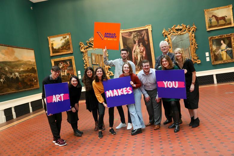 Group photo of men and women posing in an art gallery and holding colourful signs that say 'Apollo Project' and 'Art Makes You'.