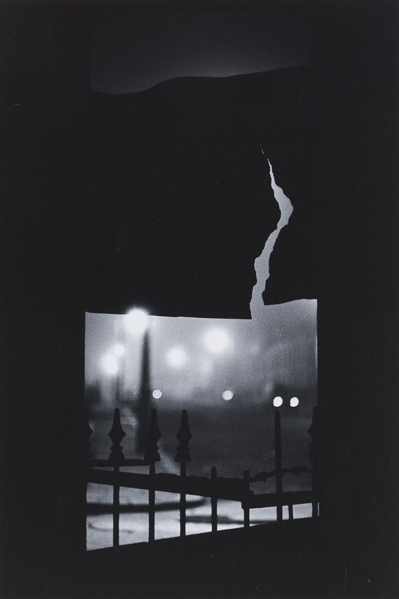 Black and white fine-art photograph of a gloomy city night scene viewed through a crack in a fence.