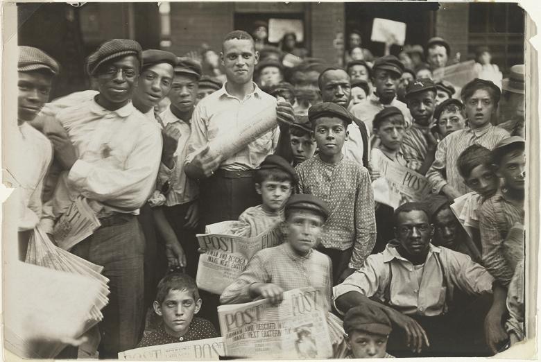 Black and white photo from 1908 showing a group of newsboys and men crowded together on the street