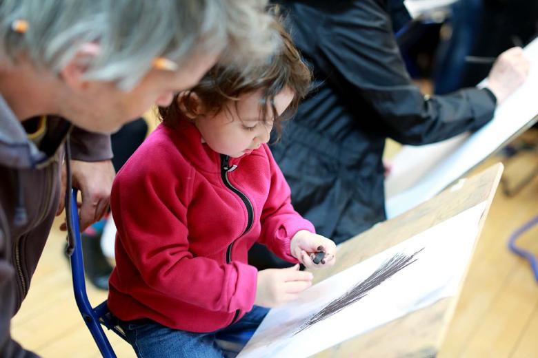 Photo of a toddler sitting on a stool and making a charcoal drawing on a board propped on their lap.