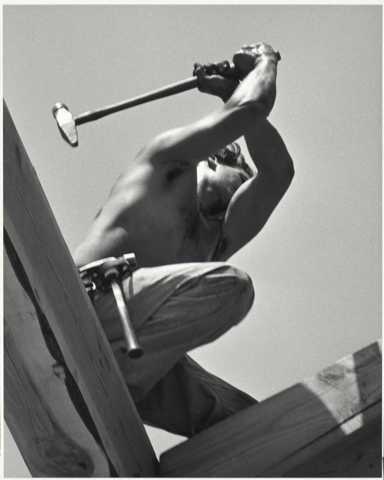 Black and white photo taken from a low angle showing a shirtless man kneeling on wood, with a hammer raised over his head