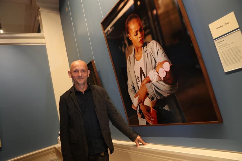 Photo of a man posing in a gallery beside a large fine-art photograph of a woman holding a child.