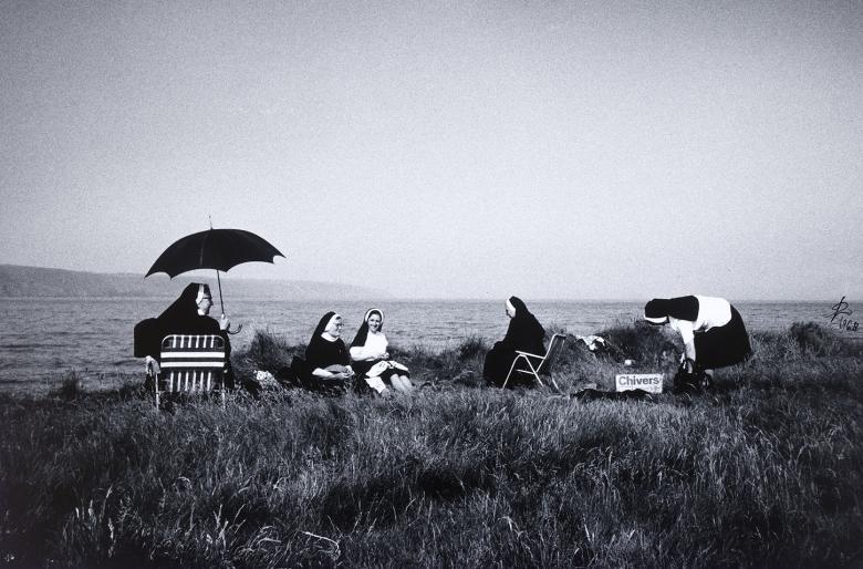 A group of nuns sit on a grassy patch overlooking the coast. Some sit on deckchairs, one with an umbrella over her head to shade her from the sun; others sit on blankets. 