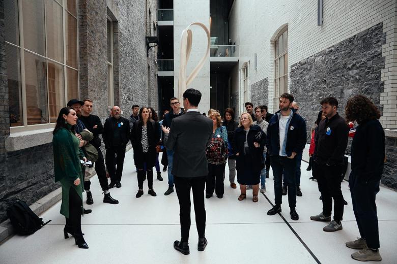 A group of people stand in a semicircle around one speaker in a light-filled courtyard space