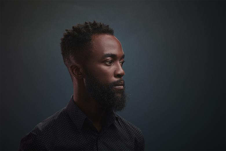 A photographic portrait of a man. He is standing slightly turned away from the viewer, and dressed in a buttoned up shirt. He gazes to the right of the frame.