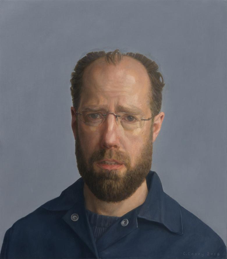The sitter looks directly at the viewer. We see his head and shoulders. He wears an open neck dark blue shirt over a dark blue t-shirt. The background is a slightly lighter shade of blue-grey.