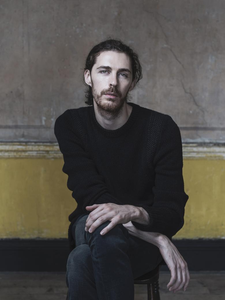 A photographic portrait. A man sits on a stool in front of a yellow and grey wall, with paint peeling. He wears a black round neck jumper, and dark jeans. His hands look as if they are in motion.