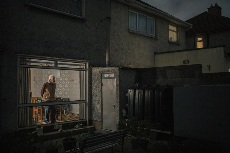 The viewer is looking in through the window of a house at night. A man stands in the illuminated window of a dining room, gazing out, hands behind his back.
