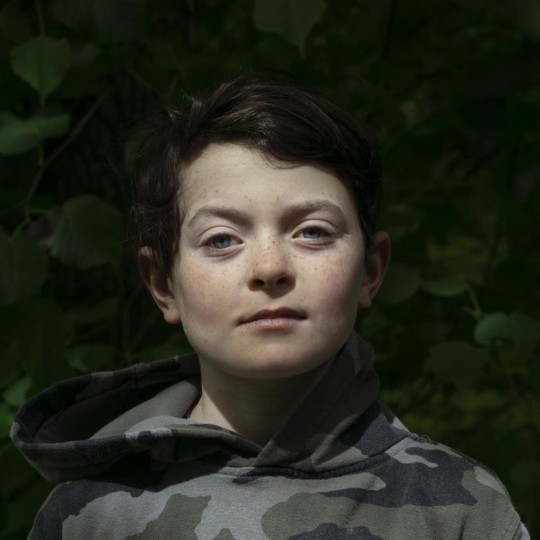 A photographic portrait of a boy. He looks directly at the camera, and is wearing a camouflage hoodie.