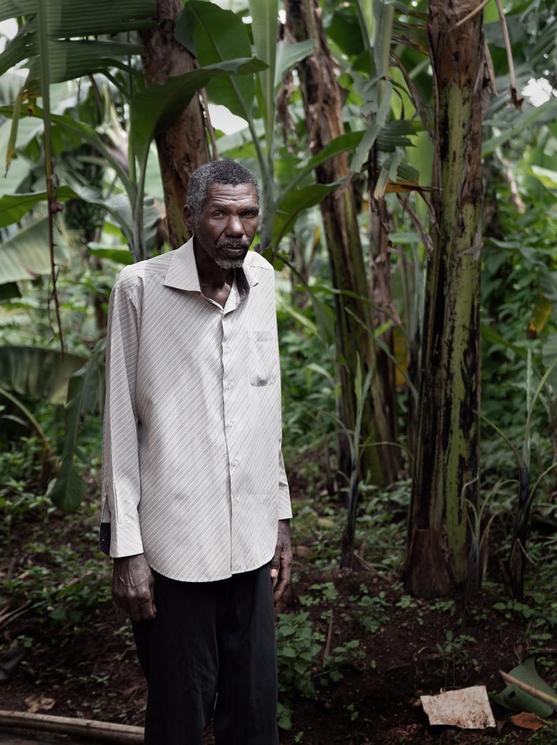 A man stands, with what thick foliage behind him. He wears a long sleeved buttoned shirt, untucked, and black pants. His arms are by his side, and he looks directly at the viewer.