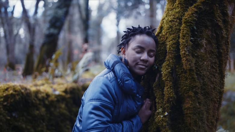 A still from a documentary film. A woman leans against a moss-covered tree, eyes closed and her ear pressed up against the trunk.
